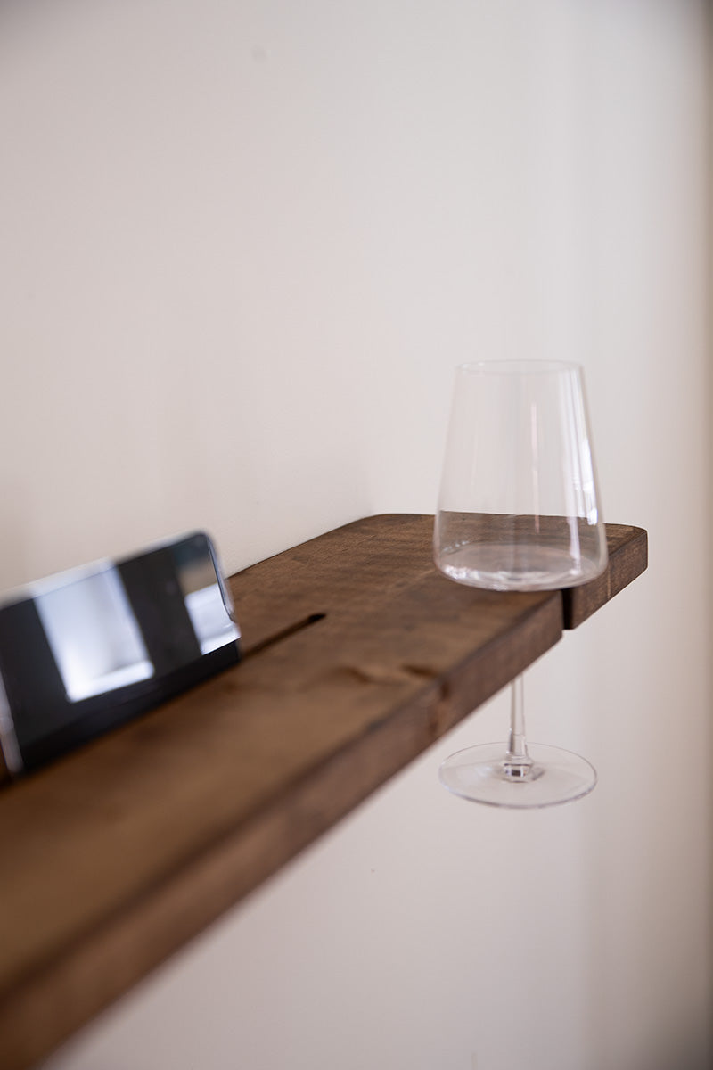 Detail van donkerbruine badplank van Baan woods met een mobiel in de sleuf en een wijnglas