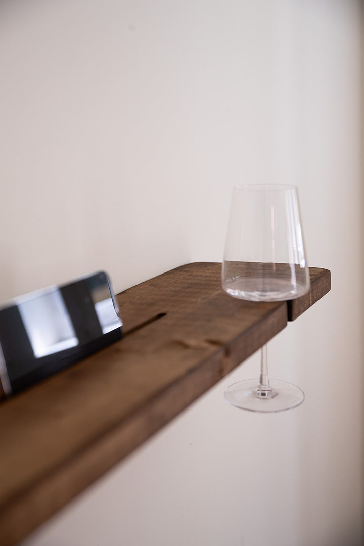 Detail van donkerbruine badplank van Baan woods met een mobiel in de sleuf en een wijnglas